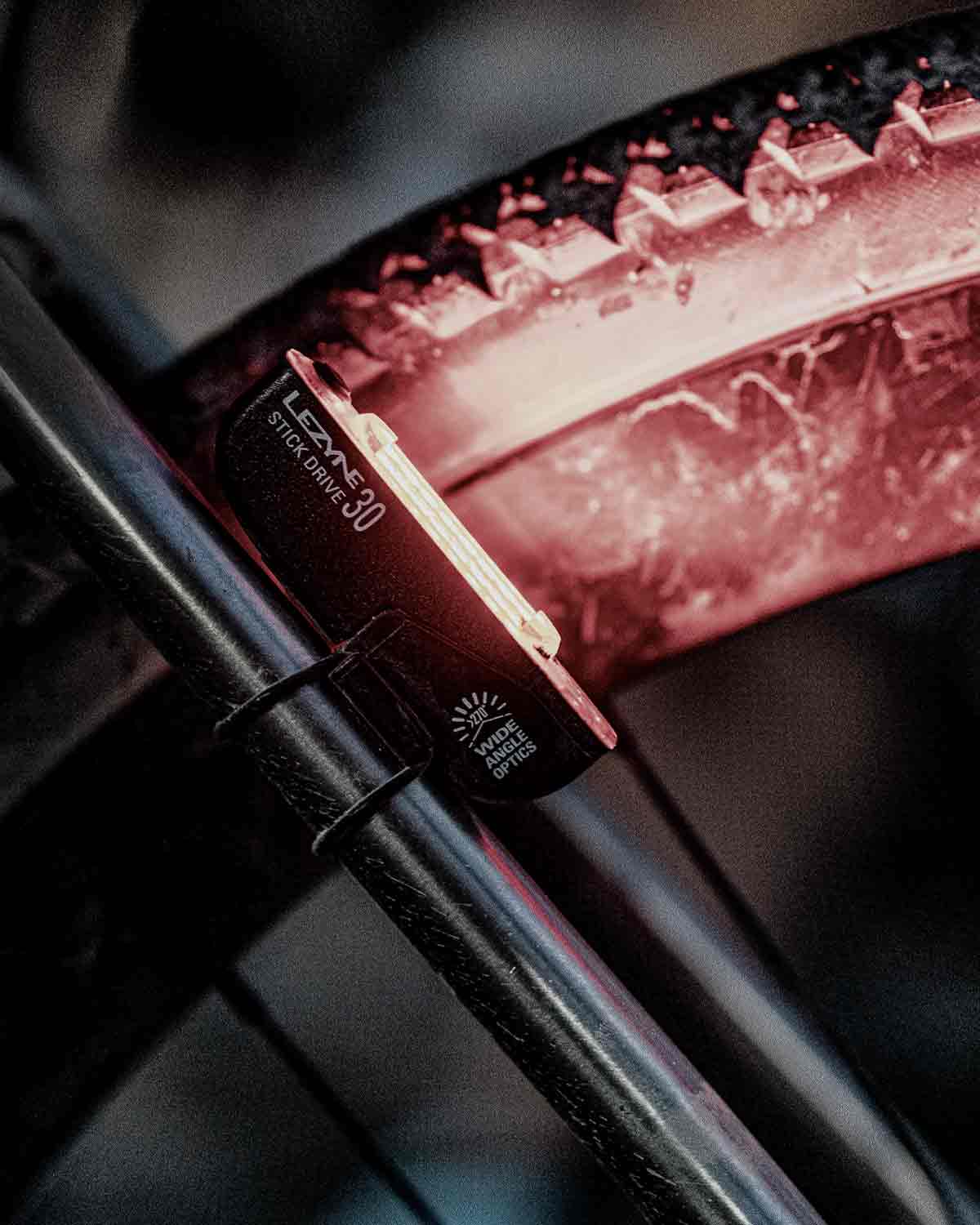 A close up of a light mounted on the rear of a bicycle that is illuminating the bicycle tire. The words Lezyne Stick Drive 30 are printed on the side of the light.