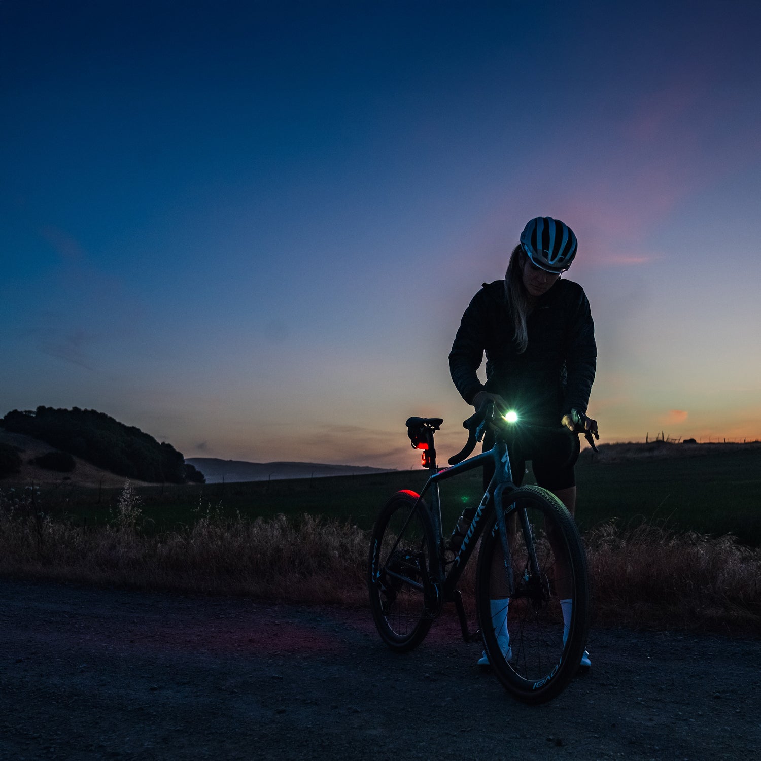 Lezyne Radar React System lifestyle scene; cyclist at road bike at dusk adjusting lights before ride, evening sky in background.