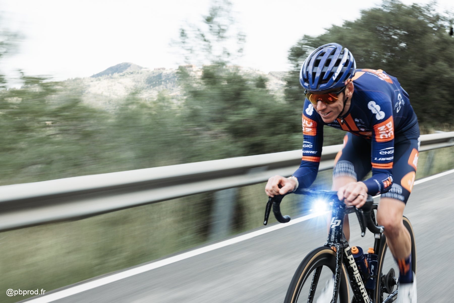 Road cyclist riding at high speed on sunlit street with bright front bike light for daytime visibility.