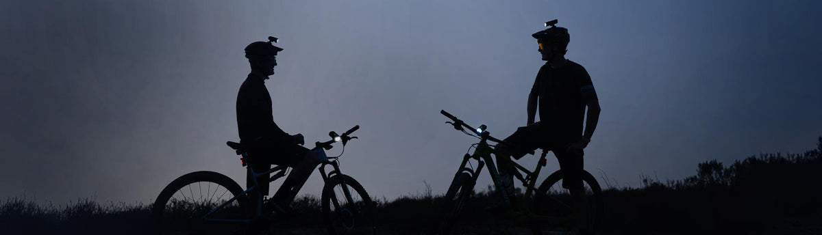 Helmet-mounted bike lights on two helmets worn by people in a dark setting, highlighting the powerful illumination and secure mounting.