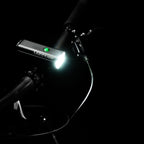 Top view of MACRO DRIVE 1400+ front LED bike light, mounted on gravel bike handlebar, showcasing a powerful light beam and green power indicator.