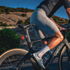 A cyclist in light gray Rapha bib shorts, white socks, and white shoes rides a black Cervelo time trial bike equipped with a sleek Lezyne Radar Pro 300 rear light on a paved road with scenic hills in the background.
