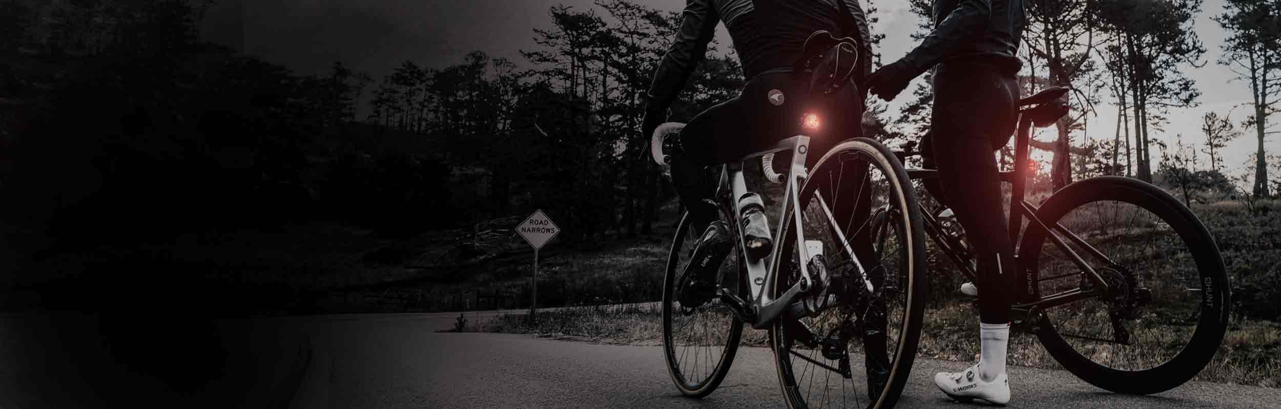 The bottom half of 2 bicycle riders stopped on the side of the road with bright rear lights mounted on their seat posts.