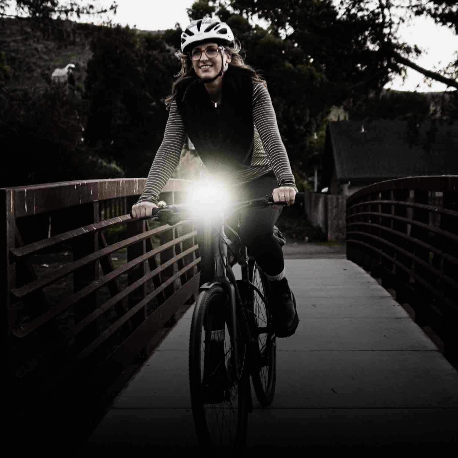A woman crossing a bridge on a bicycle with a bright light mounted on the handlebars.