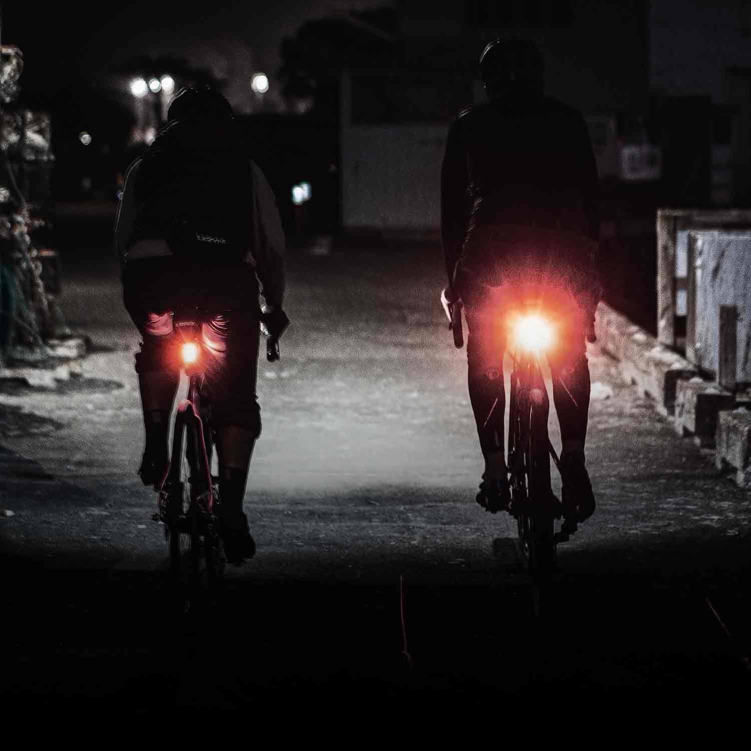 The back side of 2 people riding on a very dark road with bright red lights on the back of their bicycles and the road ahead illuminated.