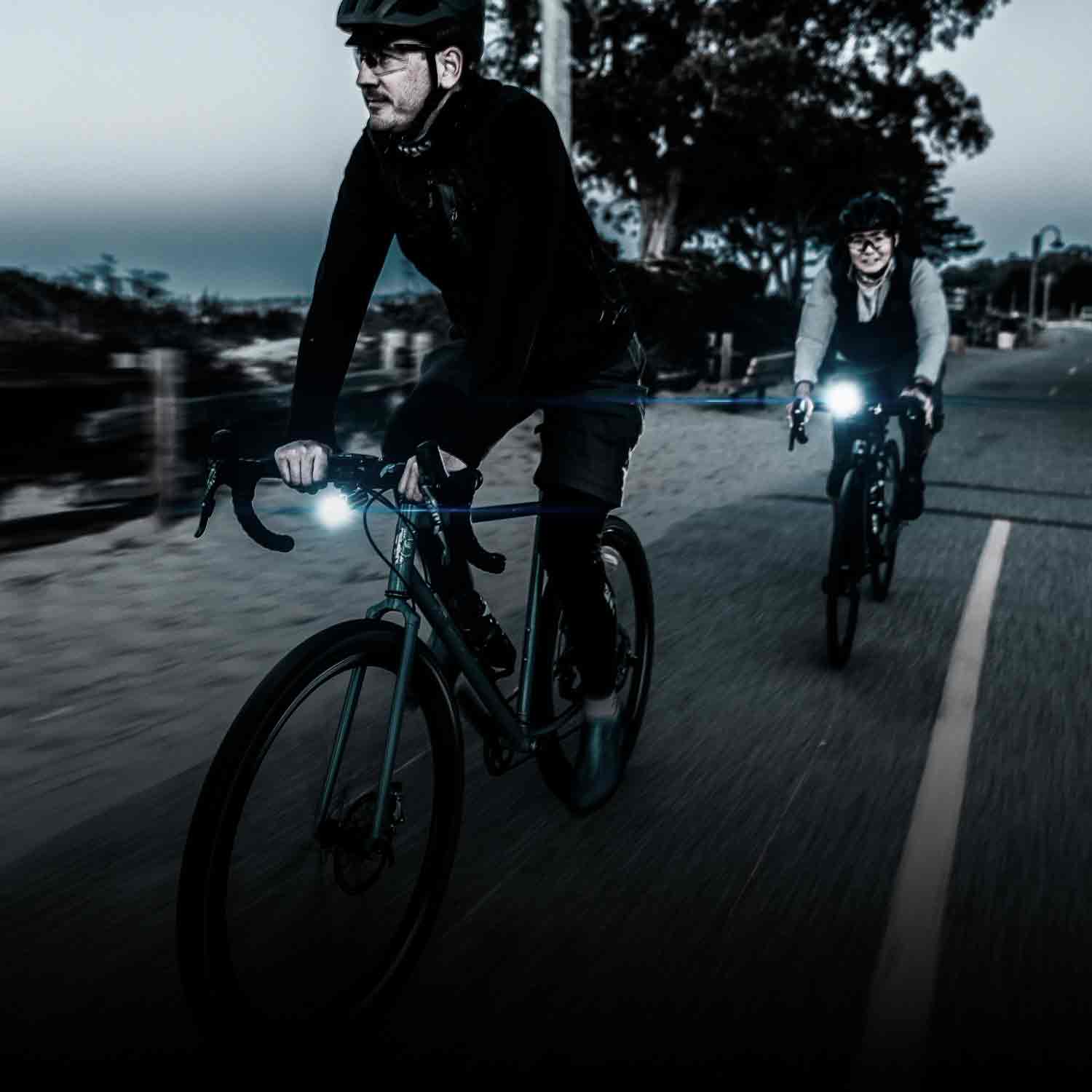 2 people riding on a road at dusk with bright lights mounted to their handlebars.