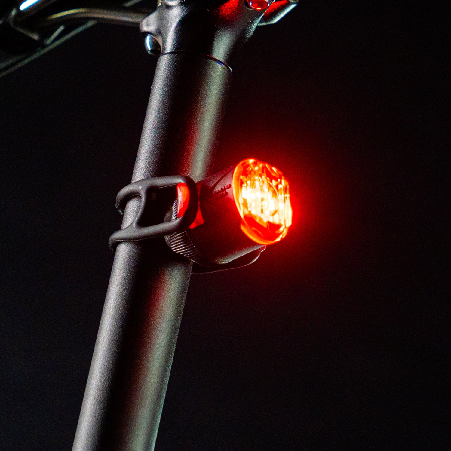 Upward side view of the circular Lezyne Femto USB Rear StVZO tail light, emitting a strong red light, mounted on the seat post with part of the saddle visible.