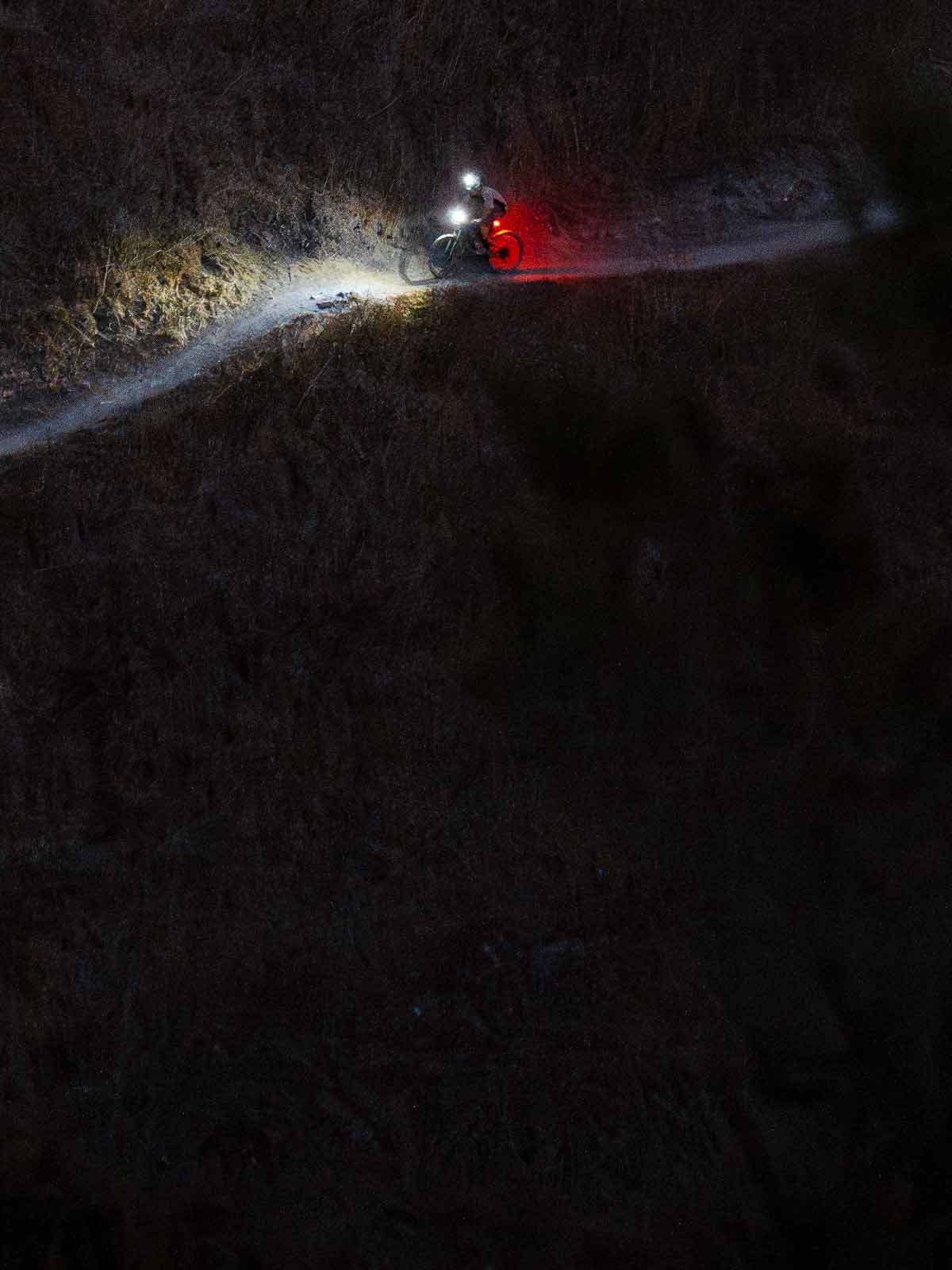 Mountain biker in the dark on the side of a hill with bright lights illuminating the trail ahead