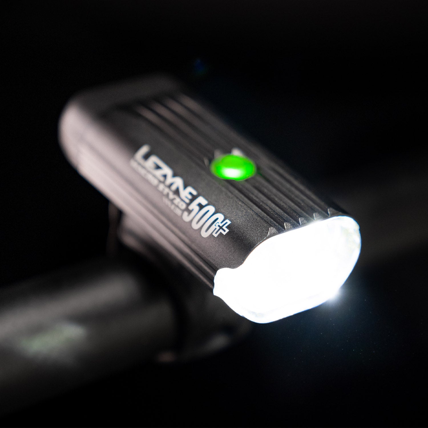 Top-down side view of the Lezyne Macro StVZO 500+ Bike Light, angled downwards to the right, with a green circular power light on top and a bright white beam, mounted on the handlebars.