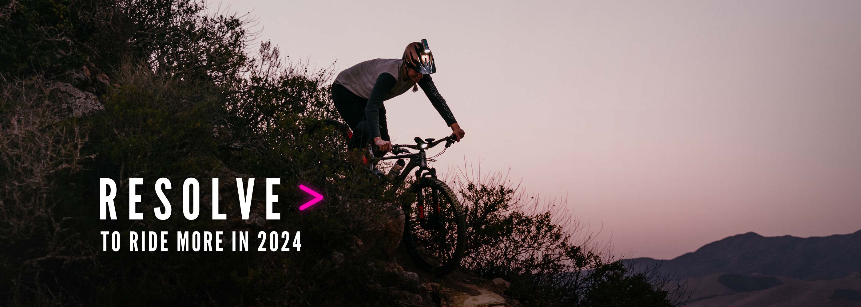 Bike rider descending down a mountain at dusk with lights with type that says 'Resolve to ride more in 2024'