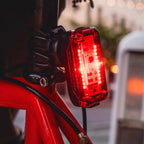 A close-up of a Radar Pro 300 bicycle tail light mounted on a red bicycle frame, with the LED light illuminated in a bright red glow against a softly blurred outdoor background.
