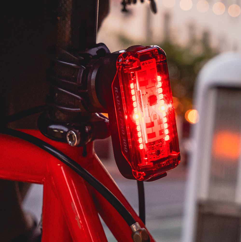 A close-up of a Radar Pro 300 bicycle tail light mounted on a red bicycle frame, with the LED light illuminated in a bright red glow against a softly blurred outdoor background.